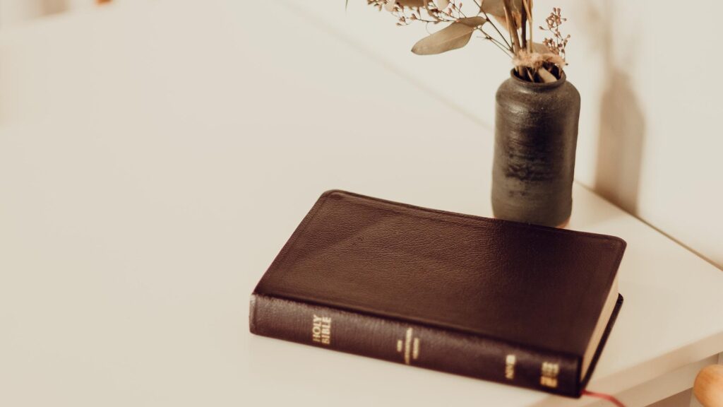 Holy bible on a table next to a vase with flowers.